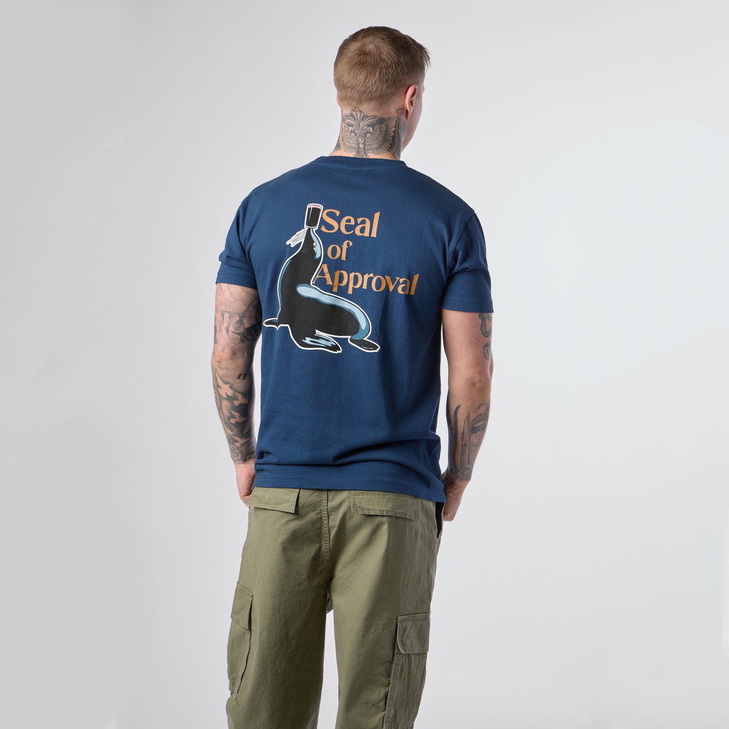 A man stands with his back to the camera, wearing the Guinness Gilroy Seal Tee – Blue from Guinness Webstore US, which features a graphic of a seal balancing a cup with the text “Seal of Approval,” paired with olive green cargo pants.