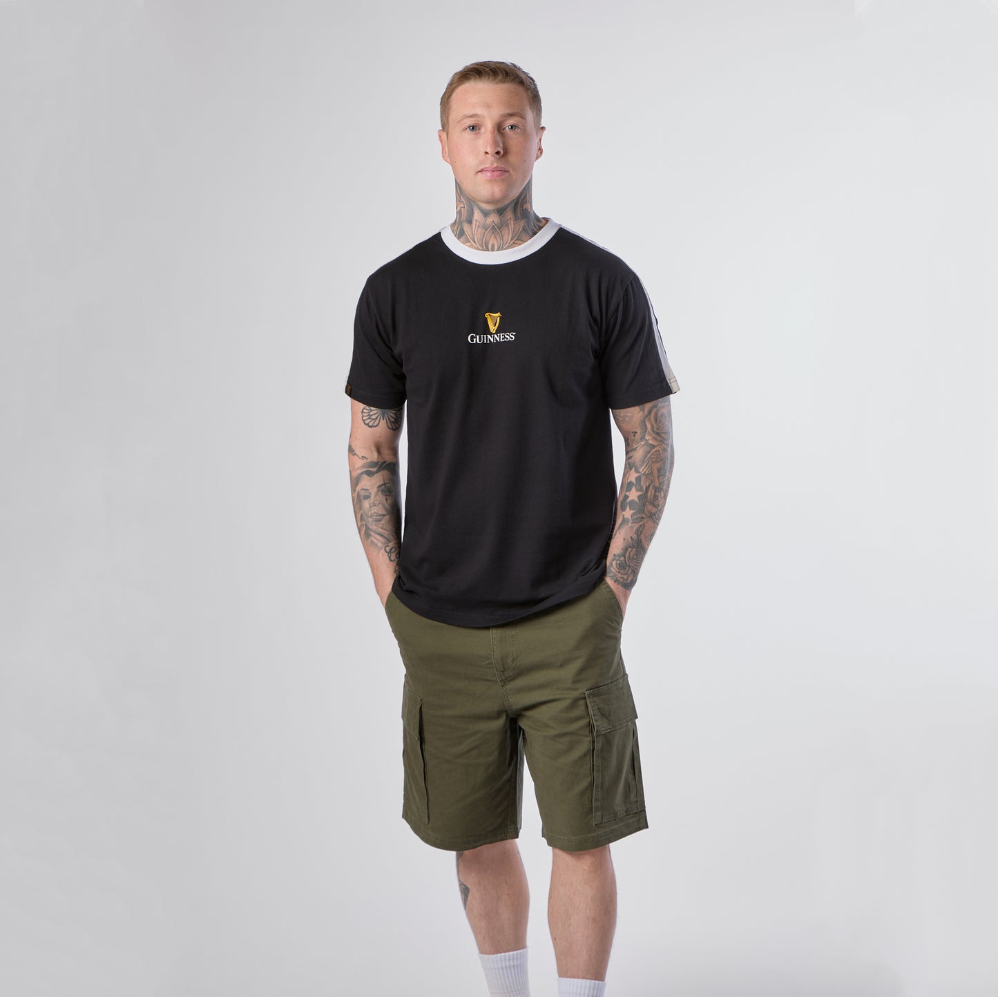 A person with tattoos is standing against a plain background, wearing a Guinness Webstore US Guinness Toucan Soccer Black Tee, olive green cargo shorts, white socks, and sneakers.