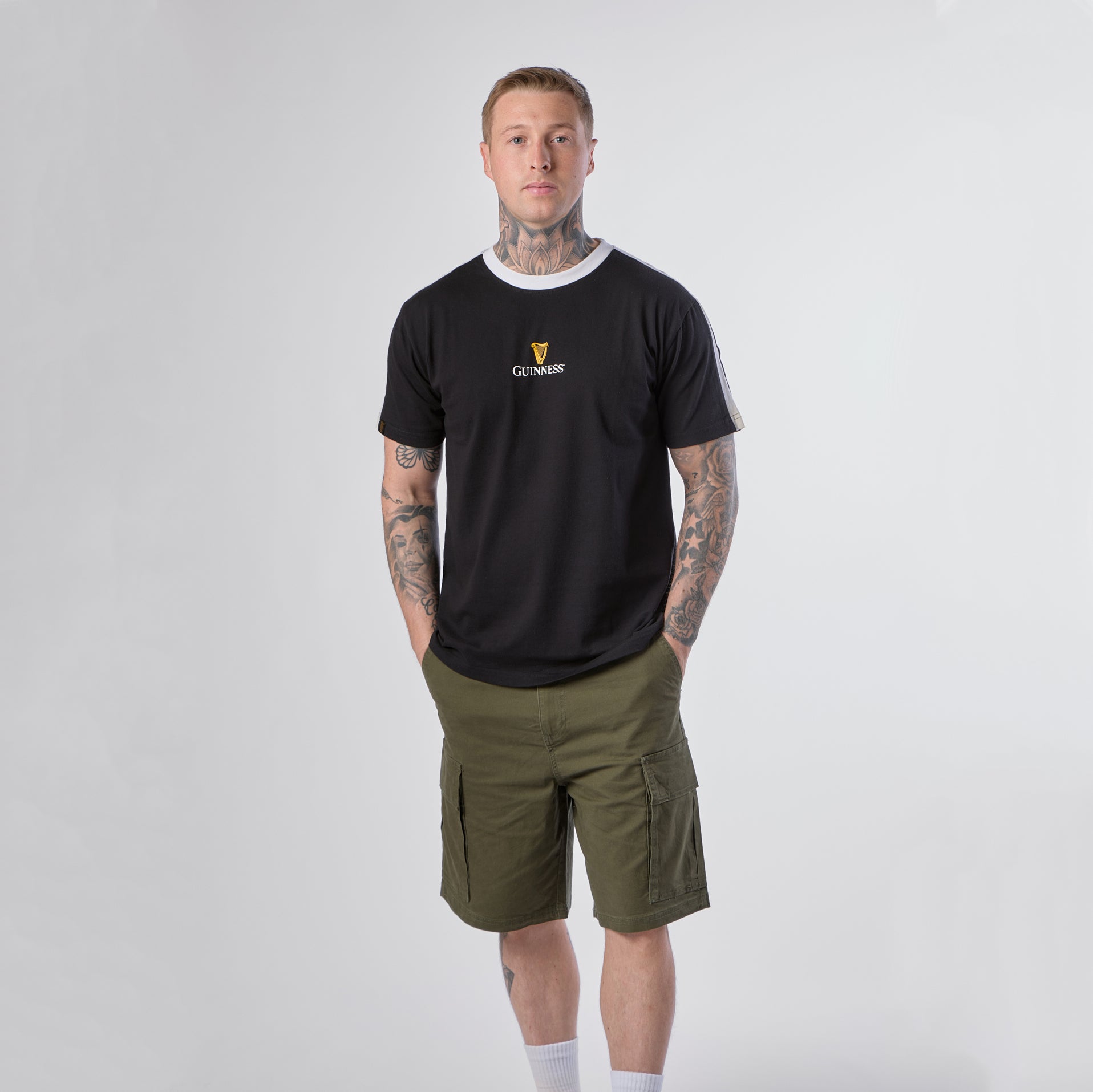 A person with tattoos is standing against a plain background, wearing a Guinness Webstore US Guinness Toucan Soccer Black Tee, olive green cargo shorts, white socks, and sneakers.