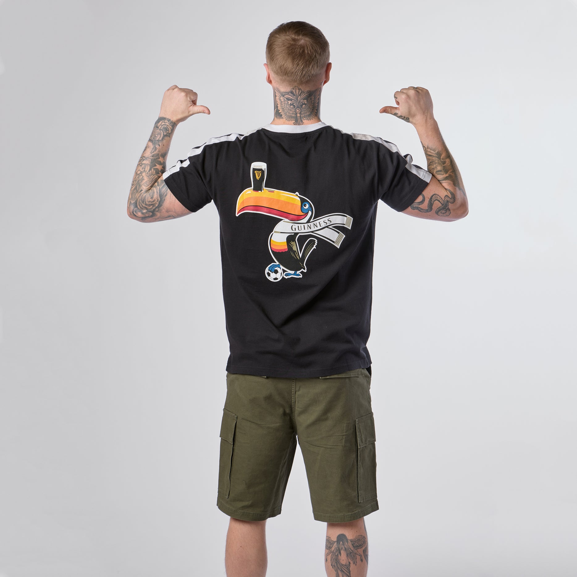 A person with tattoos, wearing the Guinness Toucan Soccer Black Tee from Guinness Webstore US—featuring a colorful toucan and “Guinness” logo—and olive cargo shorts, stands facing away with their thumbs pointing toward their back.