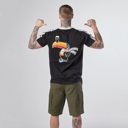 A person with tattoos, wearing the Guinness Toucan Soccer Black Tee from Guinness Webstore US—featuring a colorful toucan and “Guinness” logo—and olive cargo shorts, stands facing away with their thumbs pointing toward their back.