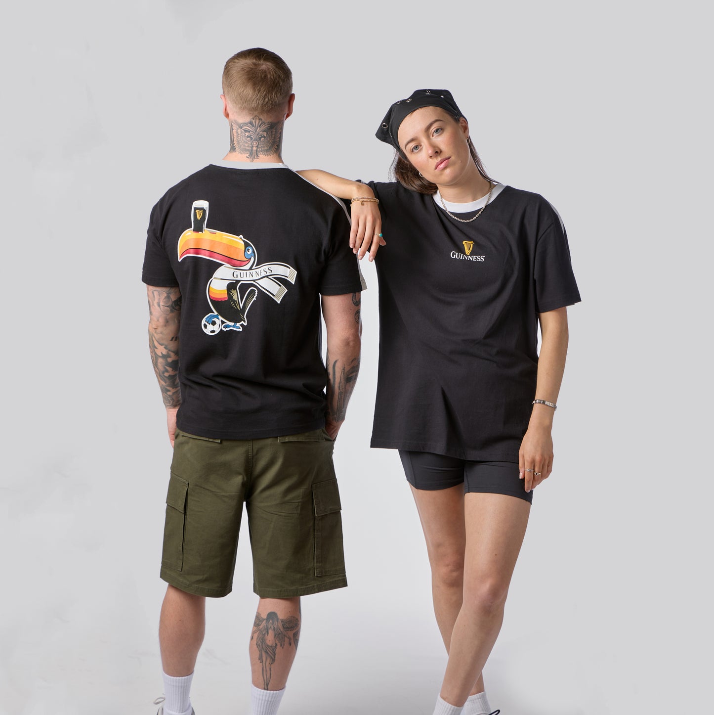 A man and a woman in casual black Guinness attire pose against a plain background; the man faces backward to display the bold graphic on the Guinness Toucan Soccer Black Tee from Guinness Webstore US, while the woman, wearing a beanie, faces forward.
