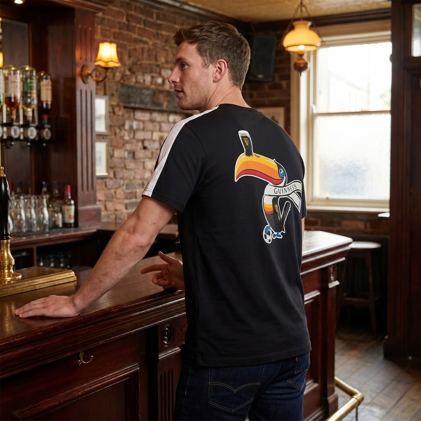 A man stands at a wooden bar in a pub, wearing the Guinness Toucan Soccer Black Tee from Guinness Webstore US, featuring a colorful toucan graphic toucan with a "Guinness" scarf, pint on its beak, and soccer ball at its feet.