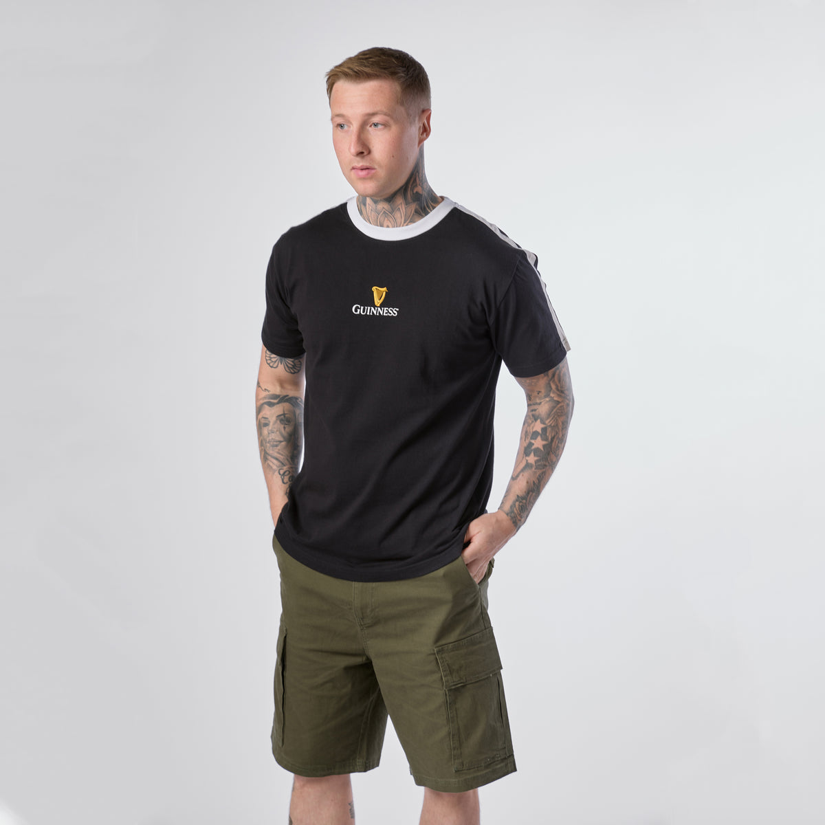 A man with tattoos wears the Guinness Toucan Soccer Black Tee from Guinness Webstore US and green cargo shorts, standing with his hands in his pockets against a plain light background.