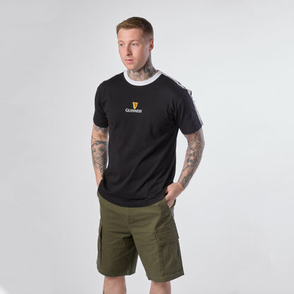 A man with tattoos wears the Guinness Toucan Soccer Black Tee from Guinness Webstore US and green cargo shorts, standing with his hands in his pockets against a plain light background.