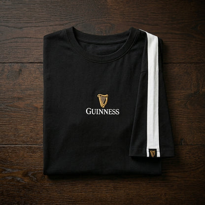 The Guinness Toucan Soccer Black Tee by Guinness Webstore US, folded on a dark wooden surface, features a white sleeve stripe, the iconic logo and harp on the chest in tribute to classic ads.