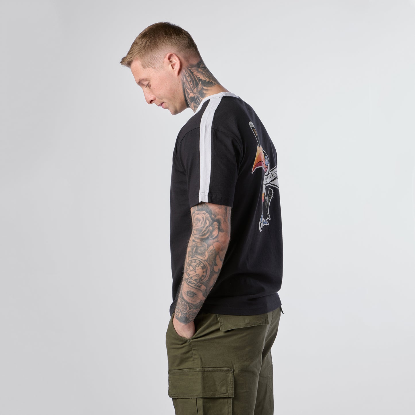 A tattooed man stands in profile, looking down with his hands in the olive cargo pockets of his pants, wearing the Guinness Toucan Soccer Black Tee from Guinness Webstore US, which features a bold graphic on the back.