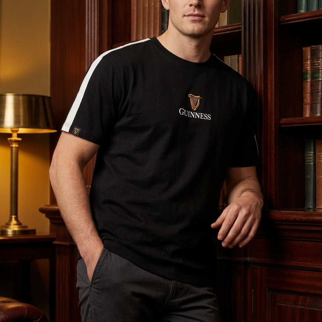 Wearing the Guinness Toucan Soccer Black Tee from Guinness Webstore US, a person stands indoors by a bookshelf with one hand in their pocket—a scene echoing the playful spirit of "My Goodness My Guinness.