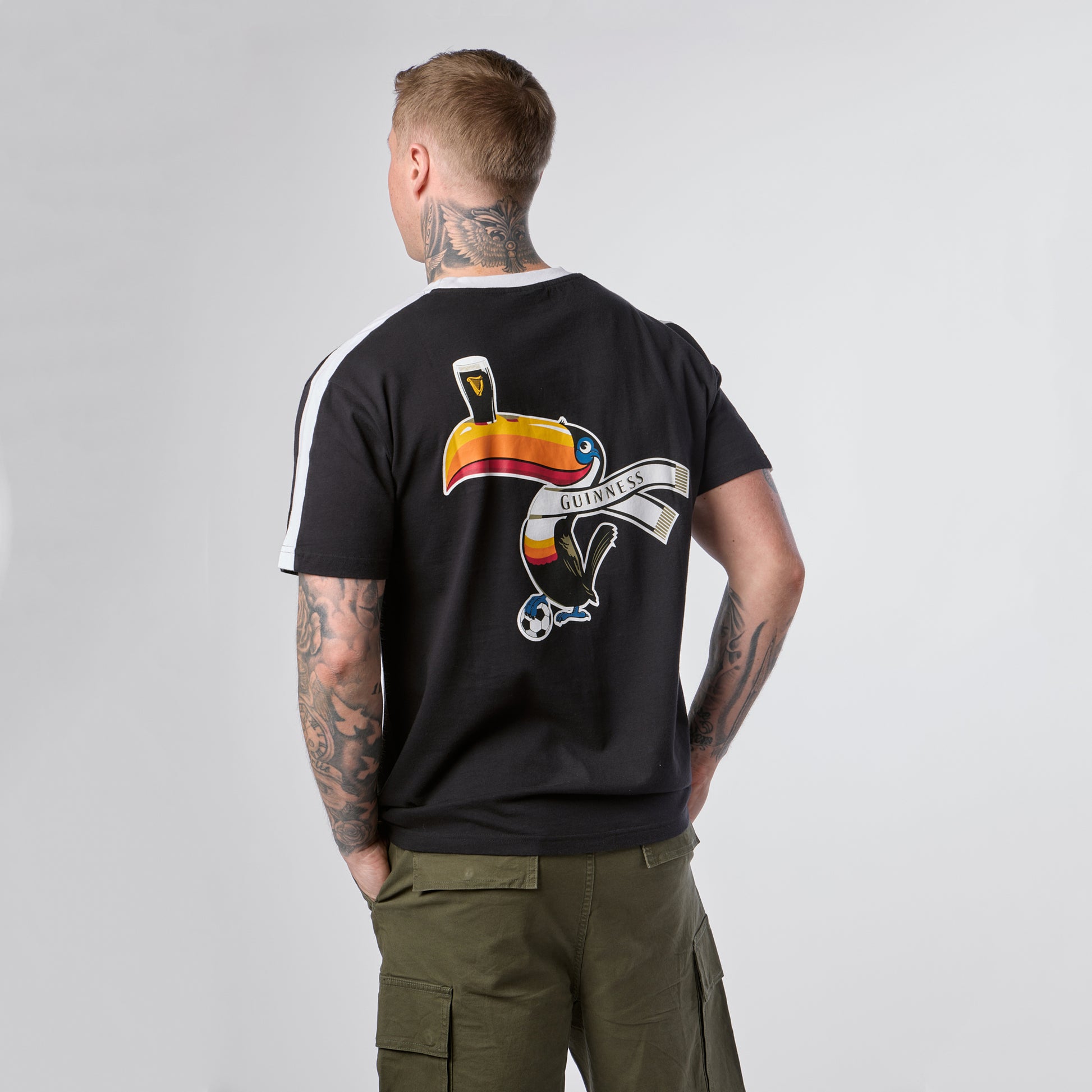 Wearing the Guinness Toucan Soccer Black Tee from Guinness Webstore US, which showcases a vibrant bird and a “Guilty Parties” banner on the back, a tattooed person stands facing away from the camera with hands in olive shorts pockets.