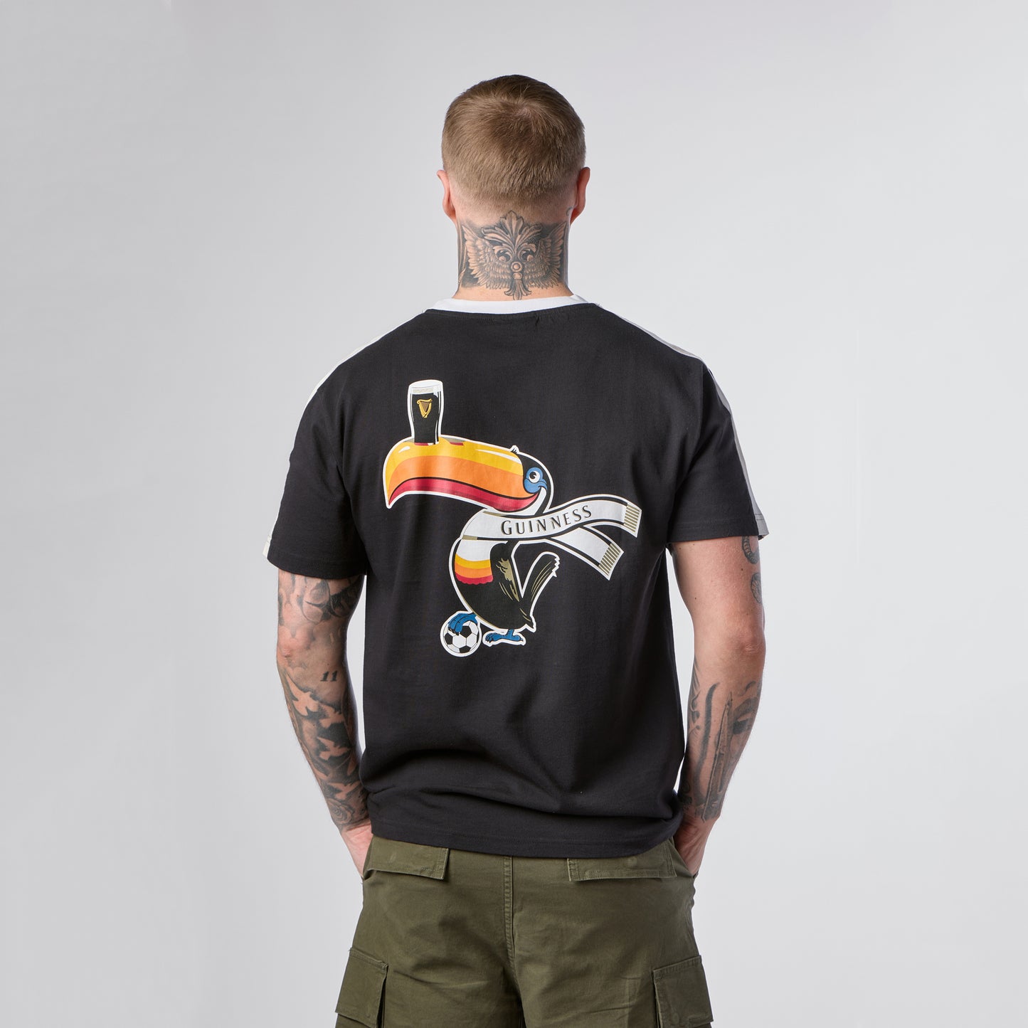 A person with tattoos on their neck and arms wears the Guinness Toucan Soccer Black Tee from Guinness Webstore US, which features a colorful toucan, a pint of Guinness, and a soccer ball graphic on the back.