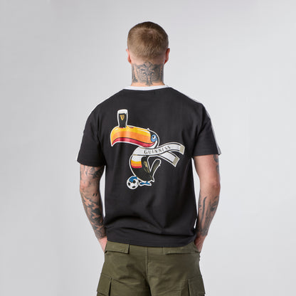 A person with tattoos on their neck and arms wears the Guinness Toucan Soccer Black Tee from Guinness Webstore US, which features a colorful toucan, a pint of Guinness, and a soccer ball graphic on the back.