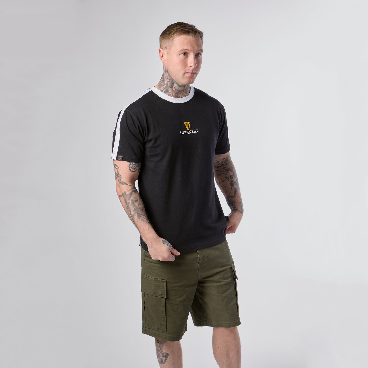 A man stands against a plain background, wearing the Guinness Webstore US Guinness Toucan Soccer Black Tee with white accents and olive green cargo shorts, showcasing tattoos on his arms and legs.