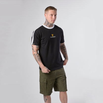 A man stands against a plain background, wearing the Guinness Webstore US Guinness Toucan Soccer Black Tee with white accents and olive green cargo shorts, showcasing tattoos on his arms and legs.