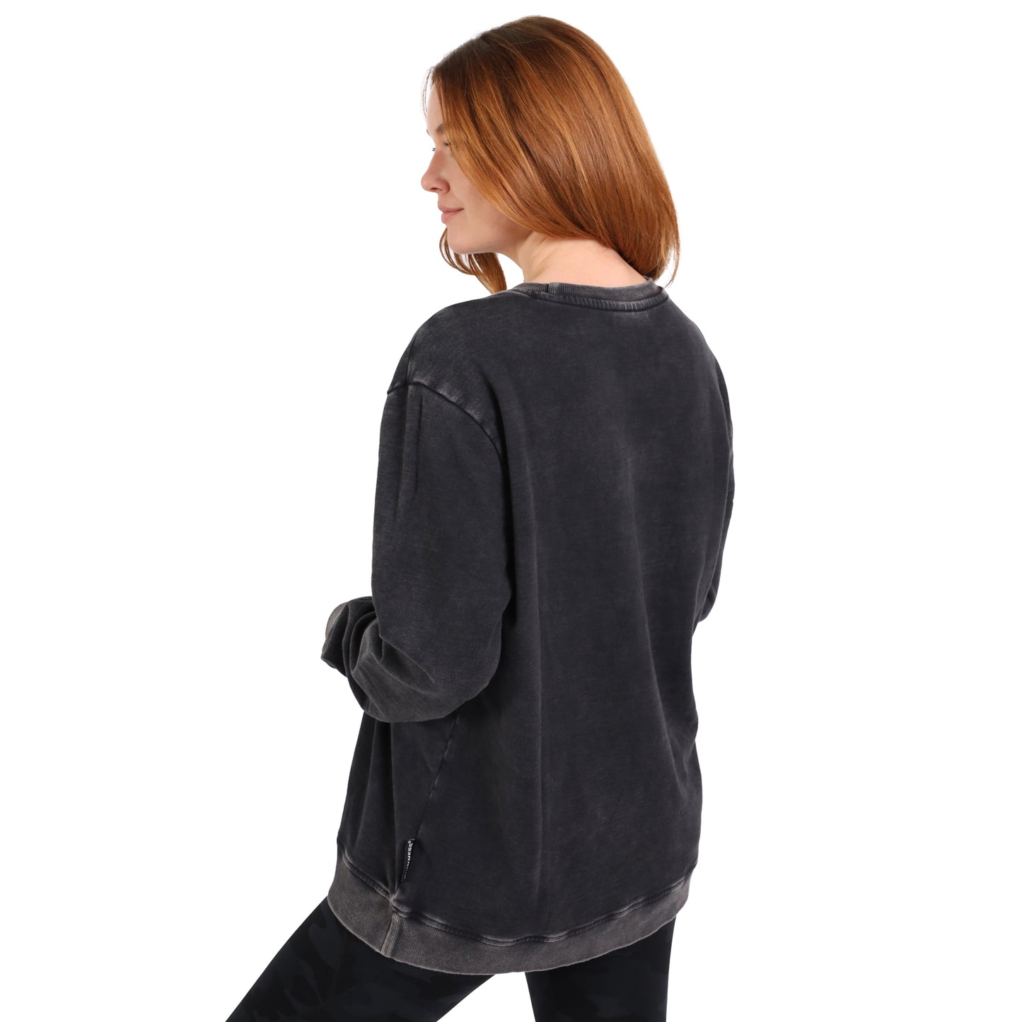 A woman with long red hair wears the Guinness Embossed Print Trademark Sweater by Guinness Webstore US and black pants, standing with her back slightly turned to the camera.