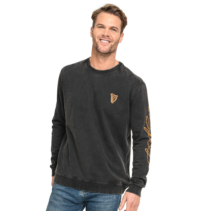 A man wearing a black Guinness Gold Harp & Signature Distressed Sweatshirt featuring a gold harp logo on the chest and gold signature text on the left sleeve.