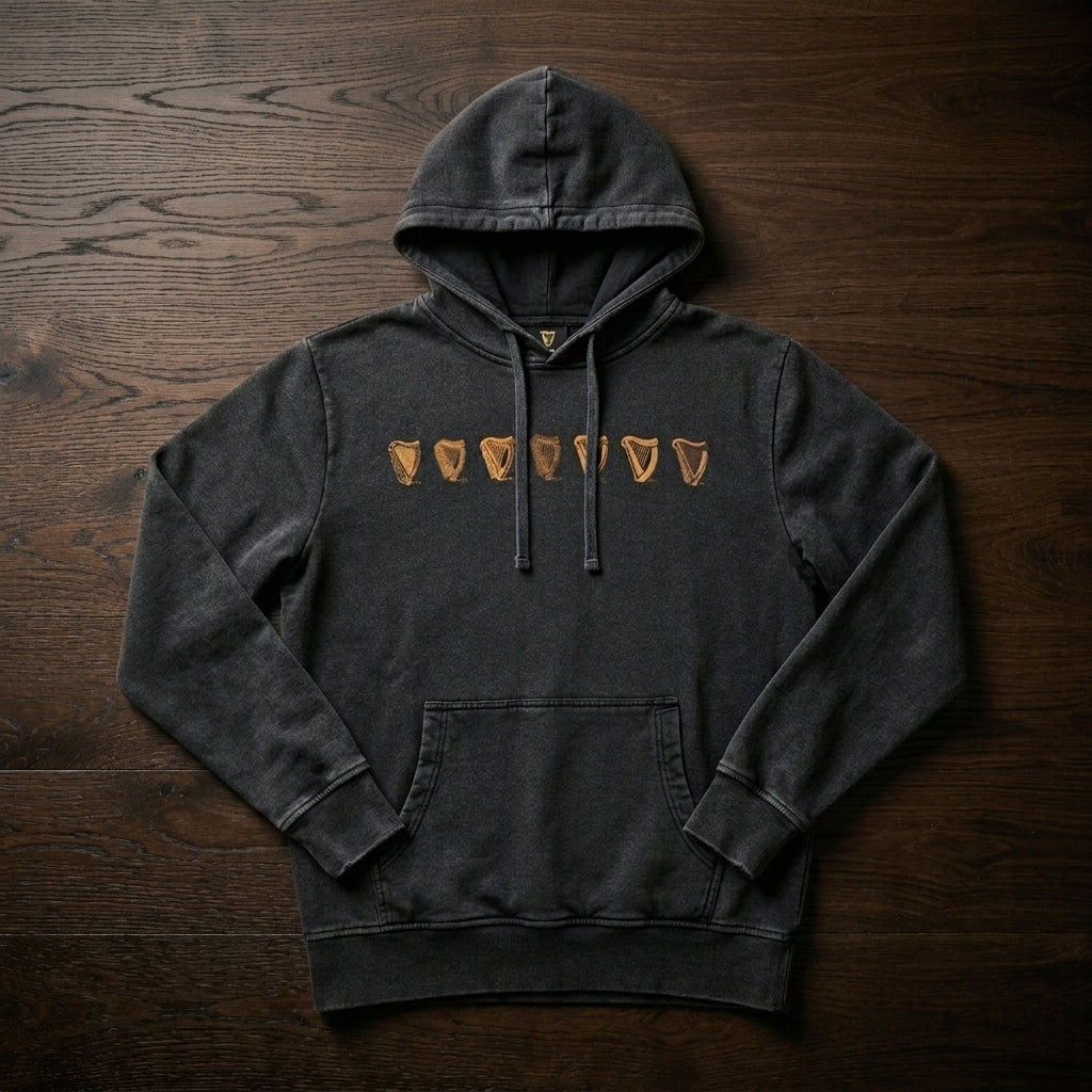 The Guinness Evolution of the Harp Distressed Hoodie – Black features six tan shield-like symbols on the chest and is crafted from dark gray cotton, shown laid flat on a dark wooden surface.