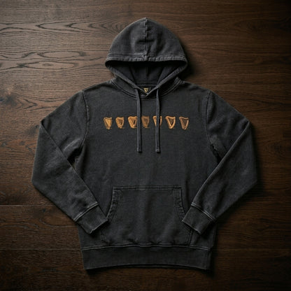 The Guinness Evolution of the Harp Distressed Hoodie – Black features six tan shield-like symbols on the chest and is crafted from dark gray cotton, shown laid flat on a dark wooden surface.