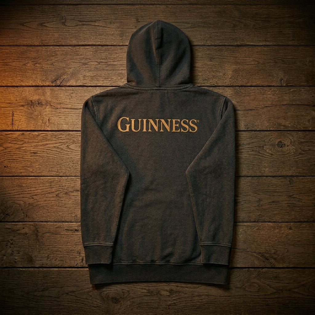 The Guinness Evolution of the Harp Distressed Hoodie in black, made from distressed cotton, features gold "Guinness" print on the back and is shown laid flat on a wooden surface.