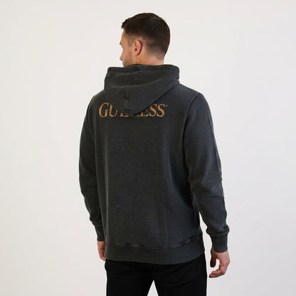 A man wears the Guinness Evolution of the Harp Distressed Hoodie in black, featuring gold "GUINNESS" lettering on the back, standing against a plain light backdrop—a subtle tribute to Guinness’s iconic symbol.