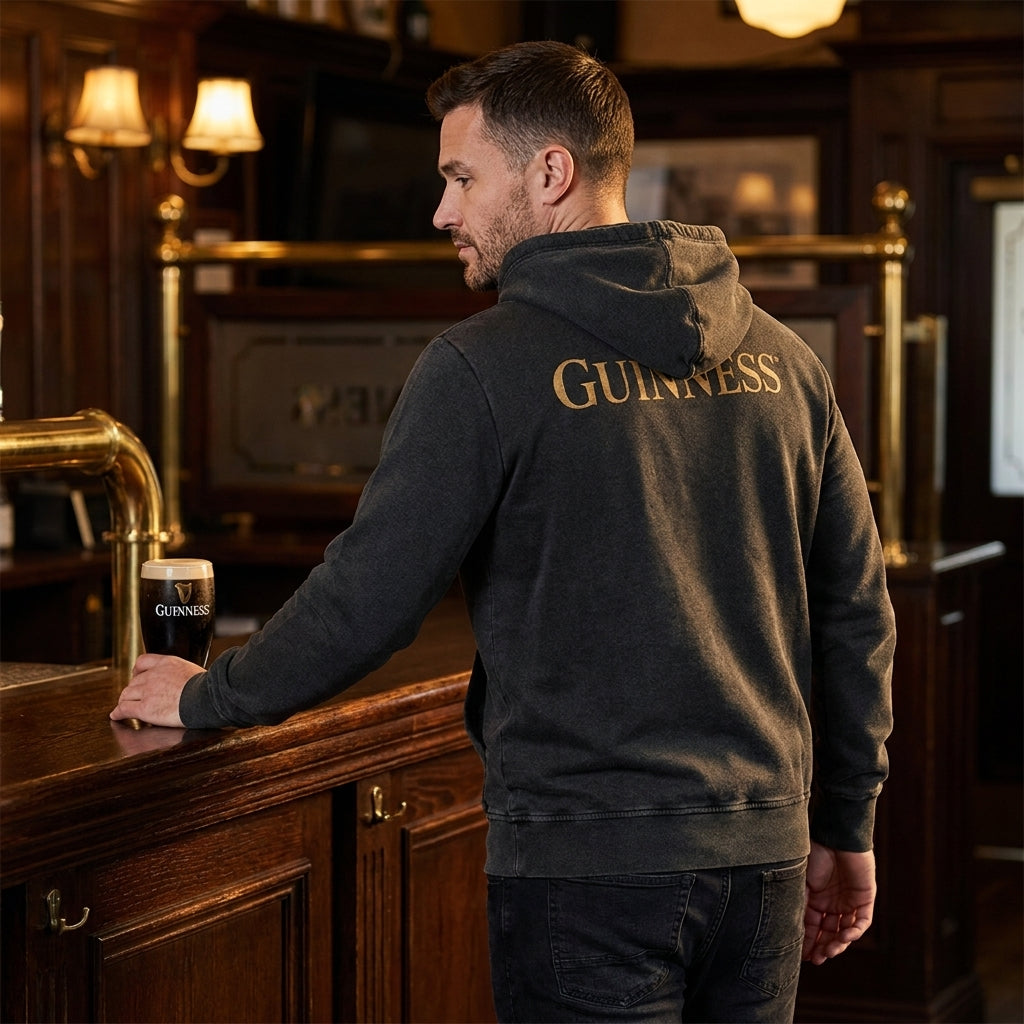 A man in a black Guinness Evolution of the Harp Distressed Hoodie stands at a wooden bar, holding a pint of Guinness, with Evolution of the Harp artwork adorning this traditional pub's walls.