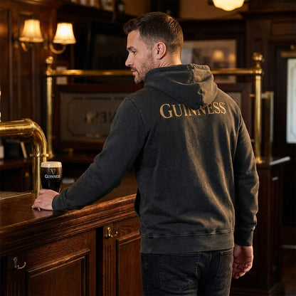 A man in a black Guinness Evolution of the Harp Distressed Hoodie stands at a wooden bar, holding a pint of Guinness, with Evolution of the Harp artwork adorning this traditional pub's walls.