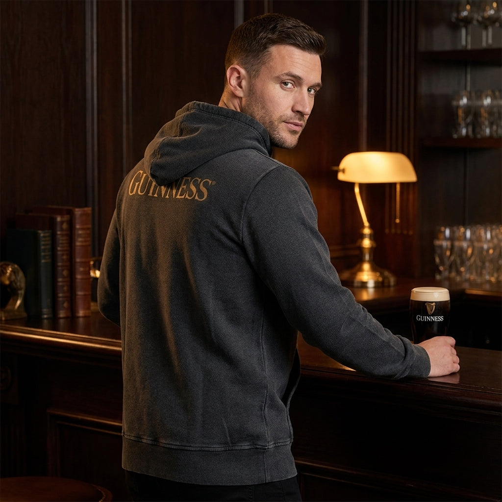 A man wearing the Guinness Evolution of the Harp Distressed Hoodie in black stands at a wooden bar, holding a pint of Guinness beer, with shelves and a lamp in the background.