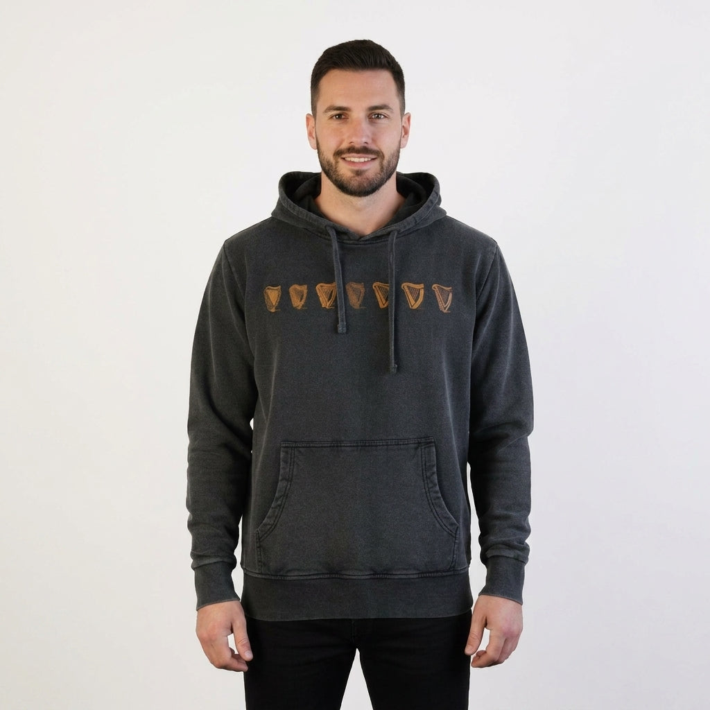 A man faces the camera in a plain white setting, wearing the Guinness Evolution of the Harp Distressed Hoodie in black, featuring five brown shield-like symbols across the chest.