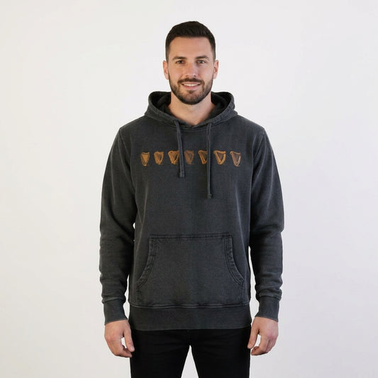 A man faces the camera in a plain white setting, wearing the Guinness Evolution of the Harp Distressed Hoodie in black, featuring five brown shield-like symbols across the chest.