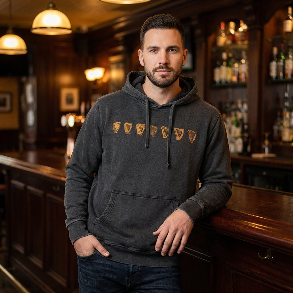 A man in a black Guinness Evolution of the Harp Distressed Hoodie stands at a wooden bar in a dimly lit pub, looking at the camera.