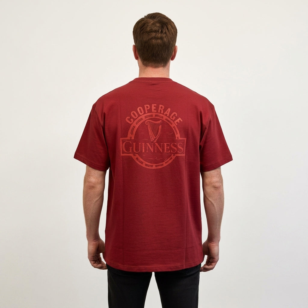 A man stands facing away, wearing the Guinness Cooperage Burnt Red Heavyweight Tee from Guinness Webstore US, featuring a circular “Guinness Cooperage” logo on the back, against a plain light background.