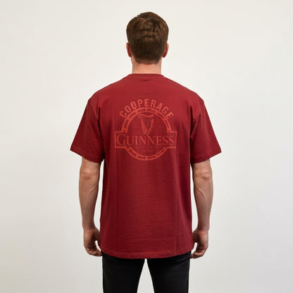 A man stands facing away, wearing the Guinness Cooperage Burnt Red Heavyweight Tee from Guinness Webstore US, featuring a circular “Guinness Cooperage” logo on the back, against a plain light background.