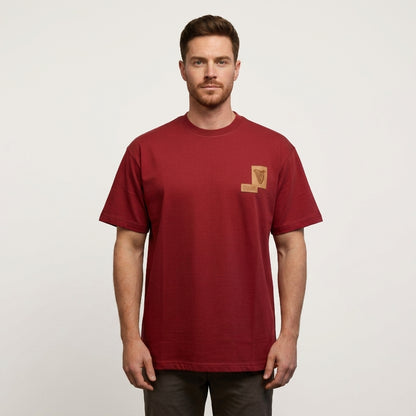A man stands facing forward against a plain white background, wearing the Guinness Cooperage Burnt Red Heavyweight Tee by Guinness Webstore US—a plain burnt red t-shirt featuring a small tan patch on the left chest.