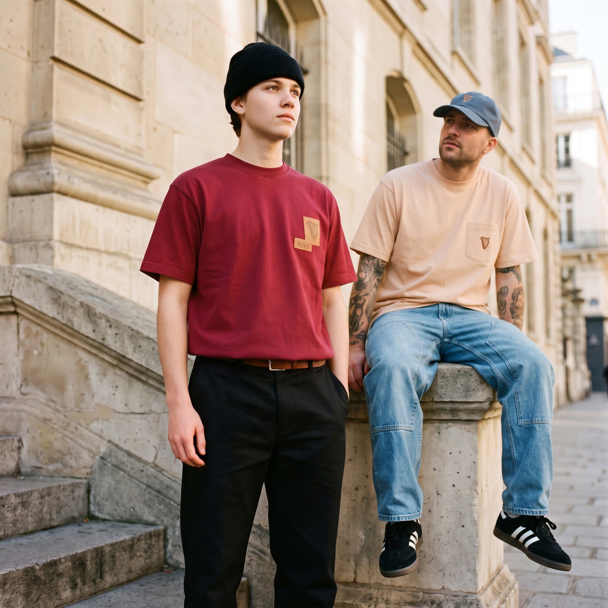 Two men in casual wear sit and stand on stone steps; one wears a maroon beanie, the other pairs jeans and a cap with the Guinness Cooperage Ivory Heavyweight Tee.