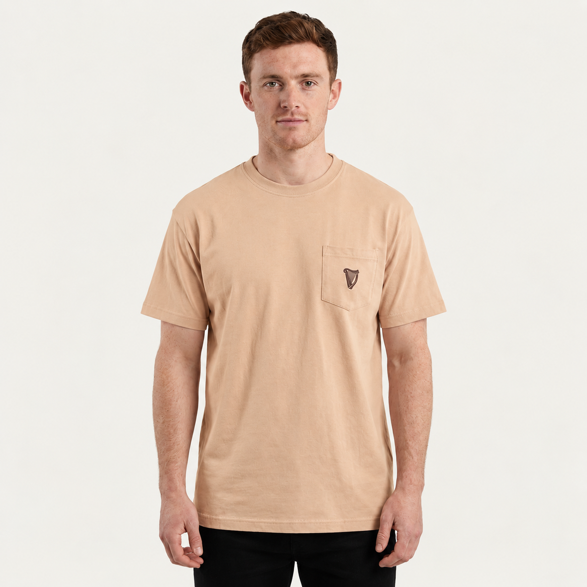 A man wears the Guinness Cooperage Ivory Heavyweight Tee—an oversized beige t-shirt with a chest pocket logo—paired with black pants against a plain white background.