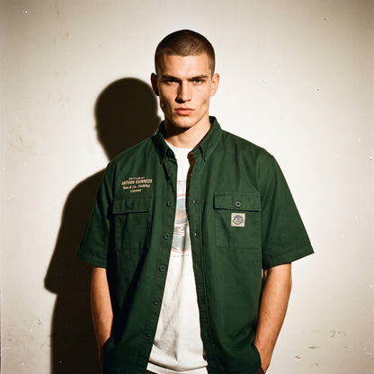 A young man with short hair stands against a plain wall wearing a green Guinness Cooperage Work Shirt from Guinness Webstore US over a white T-shirt.