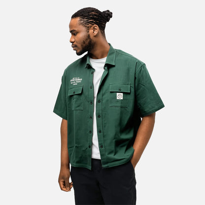 Man wears a Guinness Cooperage Work Shirt from Guinness Webstore US over a white tee with black pants, standing against a plain light background.