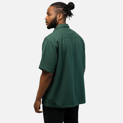 A person with braided hair in a bun wears a short-sleeve dark green Guinness Cooperage Work Shirt and black pants, standing sideways and facing away from the camera.