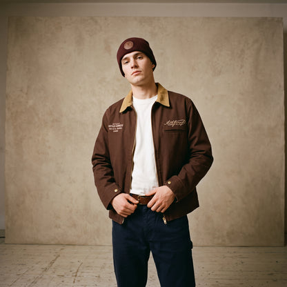 A person stands indoors against a neutral backdrop, wearing the Guinness Cooperage Work Jacket from Guinness Webstore US, paired with a white shirt, blue pants, and a maroon beanie.