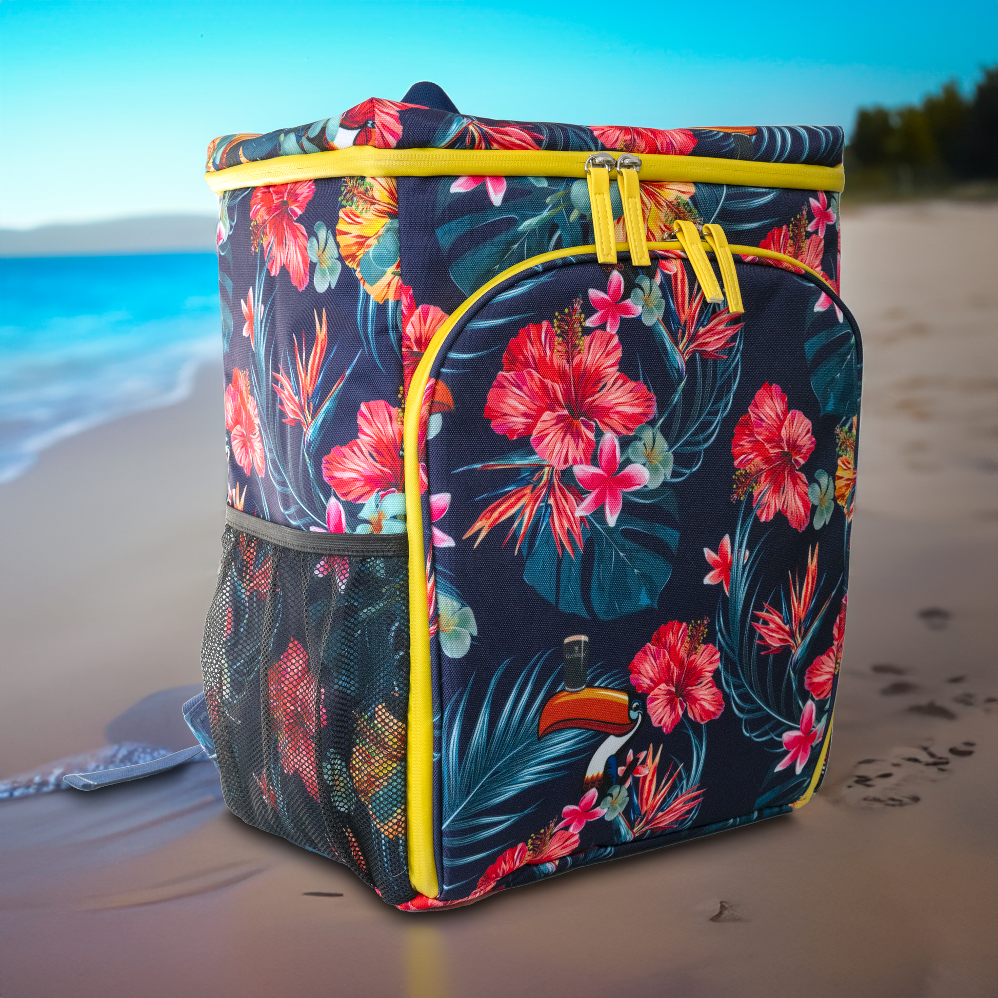 The Guinness Toucan Hawaiian Cooler Bag from the Guinness Webstore US features floral patterns, yellow zipper accents, and mesh pockets. It stands proudly on the sandy beach with the ocean in the background.