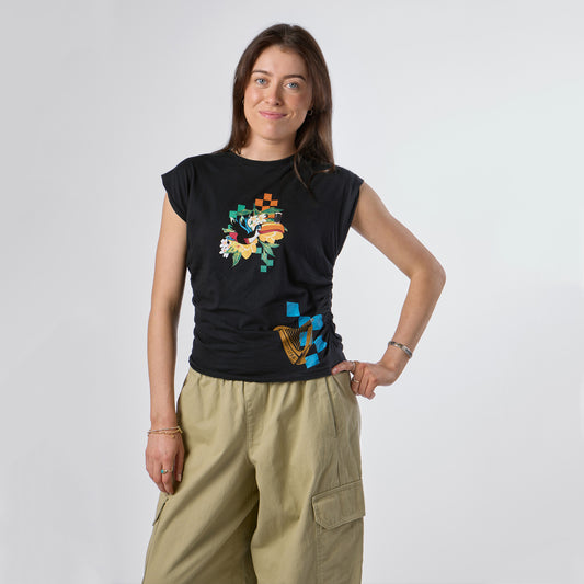 A woman stands in front of a plain background, wearing the Guinness Webstore US Guinness Toucan Ruched Ladies Black Tee with a Hawaiian-inspired graphic, paired with khaki cargo pants; she has one hand on her hip and maintains a neutral expression.