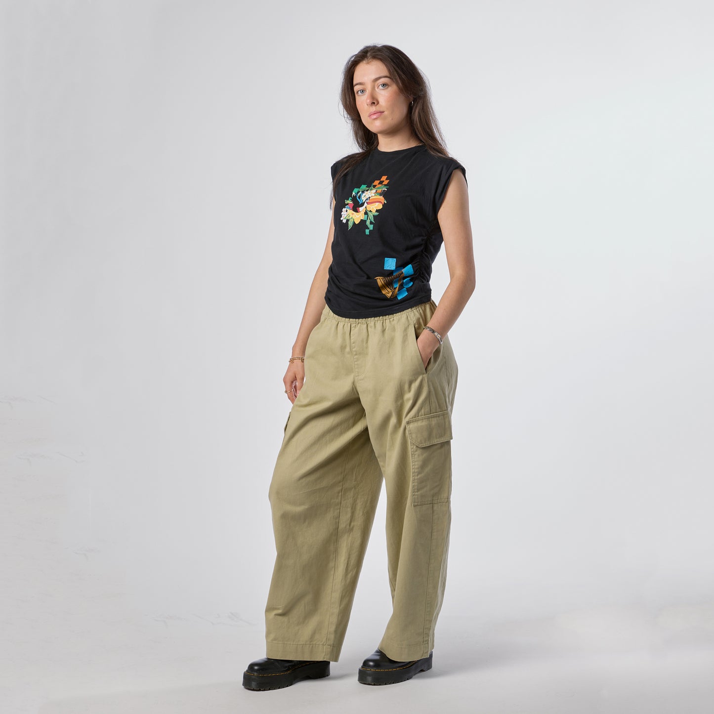 A woman stands against a plain white background, wearing the Guinness Toucan Ruched Ladies Black Tee from Guinness Webstore US, paired with khaki cargo pants and black shoes, with one hand in her pocket.