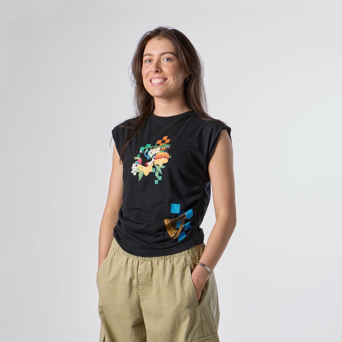 A woman stands against a plain background, smiling with her hands in her pockets, wearing light khaki pants and the Guinness Toucan Ruched Ladies Black Tee from Guinness Webstore US, which features a vibrant Hawaiian-inspired graphic.