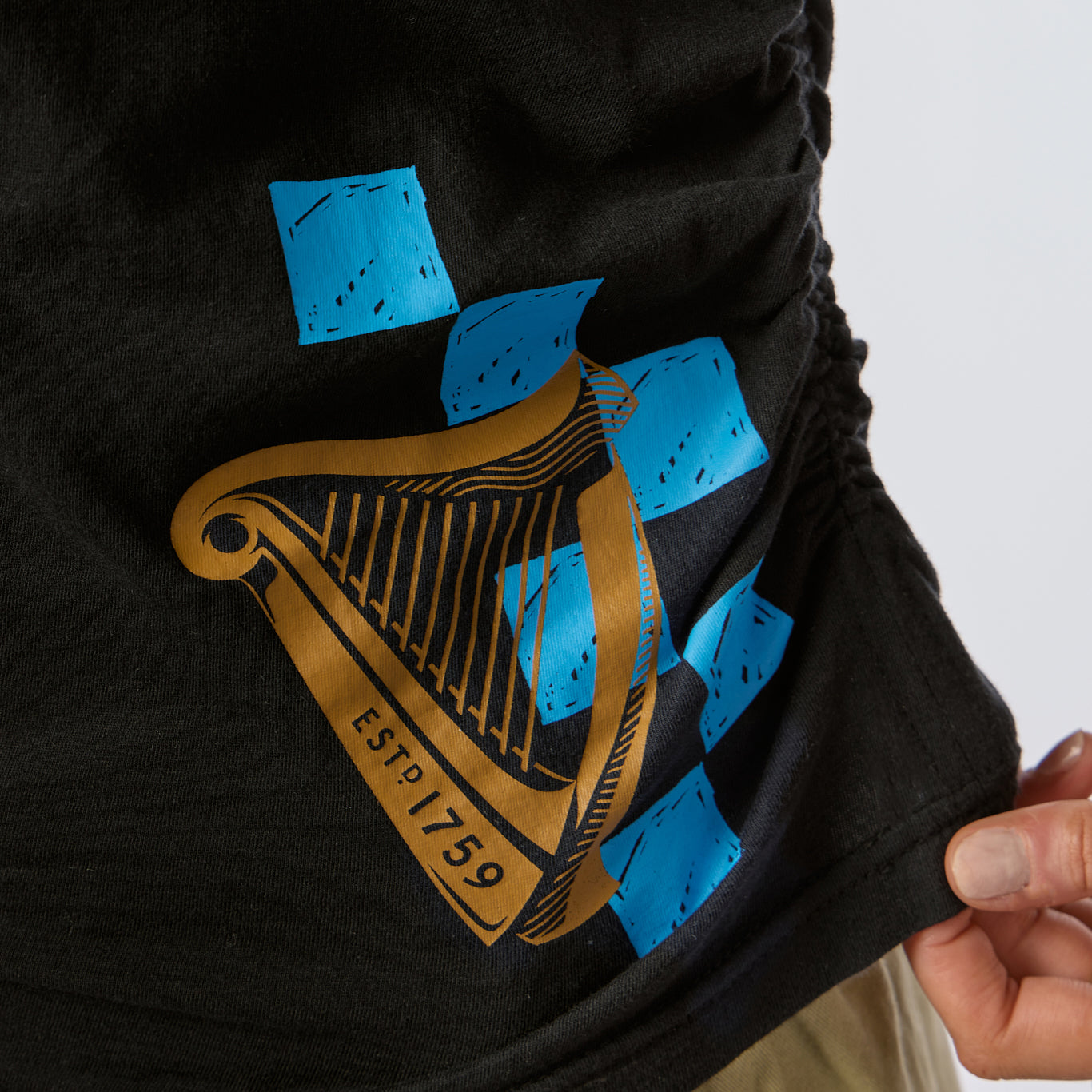 A close-up of the Guinness Toucan Ruched Ladies Black Tee from Guinness Webstore US shows a gold harp illustration labeled “ESTD 1759” overlaid with several blue geometric shapes. This tee combines classic style with modern design elements for a truly unique look.
