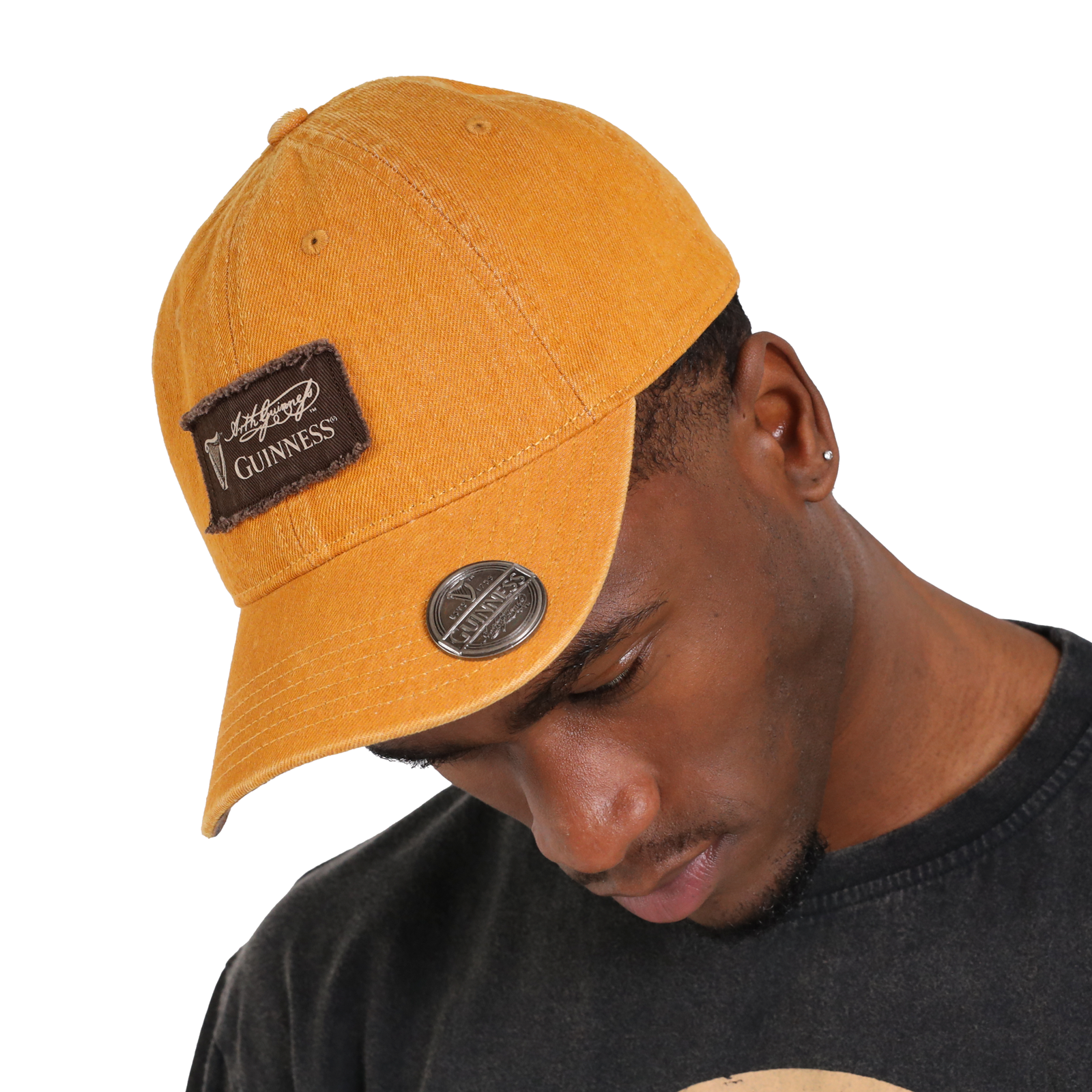 A person wearing a Guinness Mustard Bottle Opener Cap featuring the iconic Guinness Harp Logo and a decorative badge looks down, dressed in a dark t-shirt.