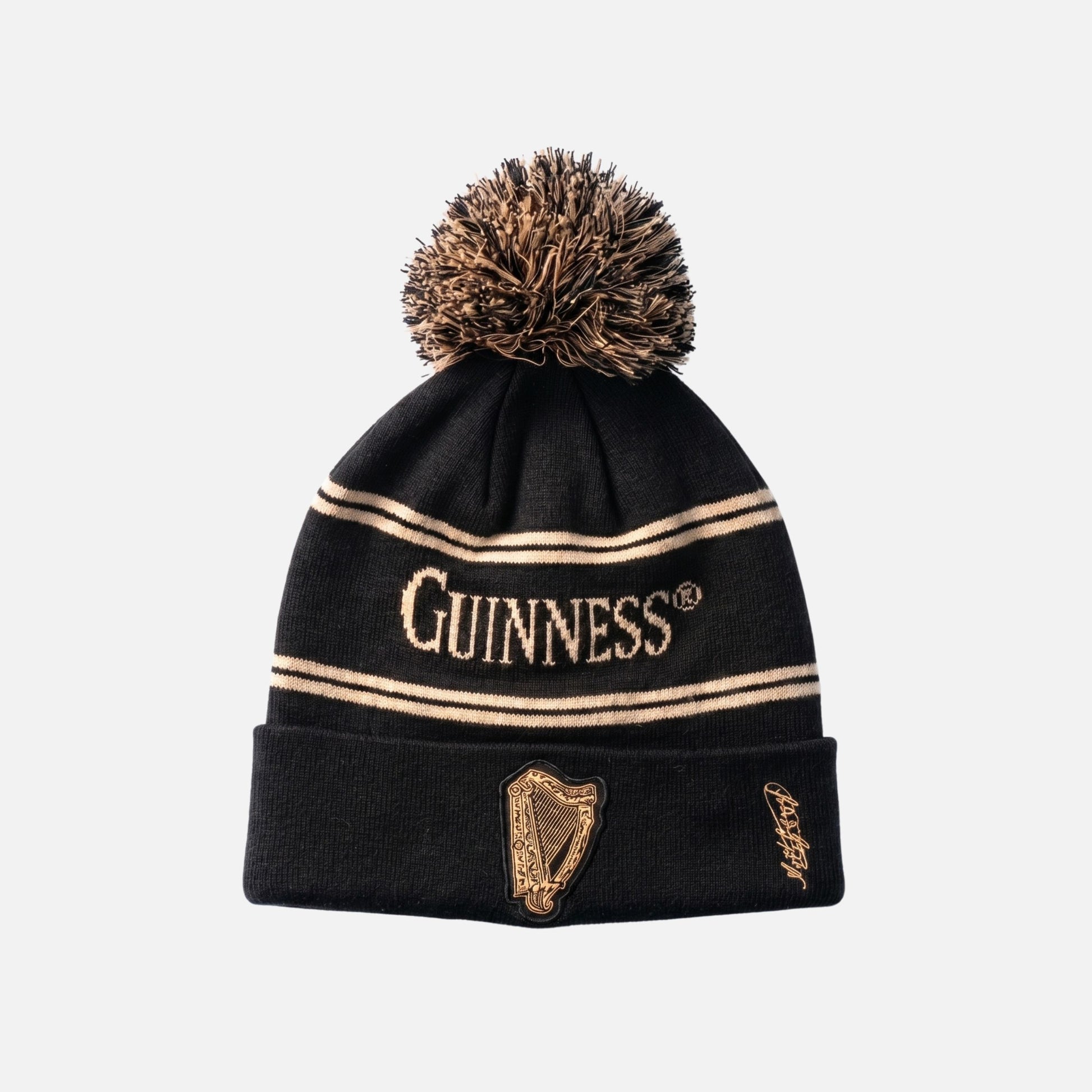 The Guinness Black & Gold Sports Beanie – Vintage Harp Knit features tan stripes, a large pom-pom, "Guinness" text, a vintage harp design, and a signature on the folded brim.