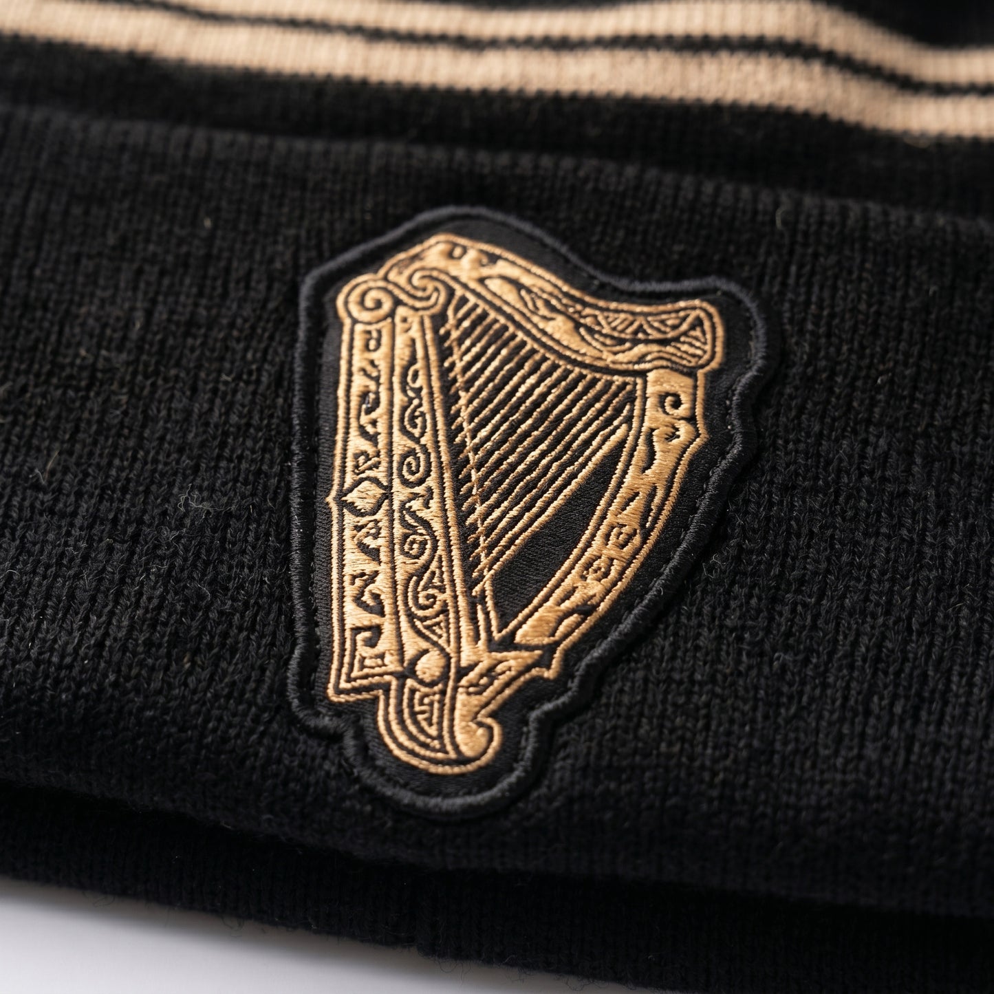 Close-up of the Guinness Black & Gold Sports Beanie – Vintage Harp Knit, a black beanie by Guinness featuring a gold embroidered vintage harp patch and beige stripes at the top.