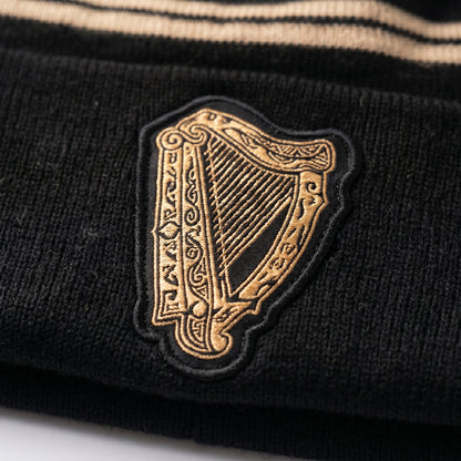 Close-up of the Guinness Black & Gold Sports Beanie – Vintage Harp Knit, a black beanie by Guinness featuring a gold embroidered vintage harp patch and beige stripes at the top.