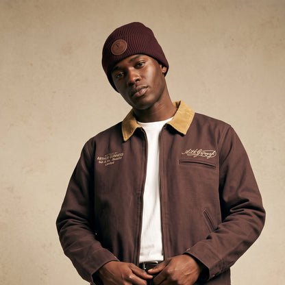 A man in a brown jacket and a Guinness Cooperage Brown Knitted Beanie by Guinness Webstore US stands against a neutral background, looking at the camera.