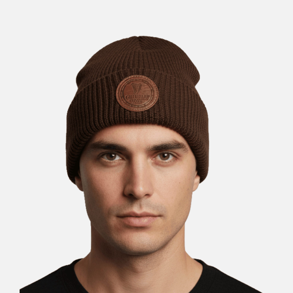 A man wears a Guinness Cooperage Brown Knitted Beanie with a round leather patch, facing the camera against a plain white background.