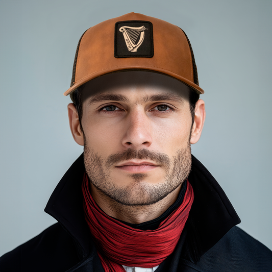 A man with a beard, wearing a Guinness Brown Leather Trucker Cap from the Guinness Webstore US featuring the harp logo, complemented by a black coat and red scarf, looks directly at the camera.